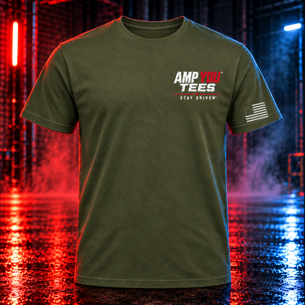 An olive green short-sleeve t-shirt with a graphic on the left chest reading 'AMP TEES' in white and red, and 'STAY DRIVEN' underneath, along with a distressed American flag graphic on the right sleeve. The shirt is displayed against a moody, wet industrial background with red and blue neon lights reflecting on the wet floor.