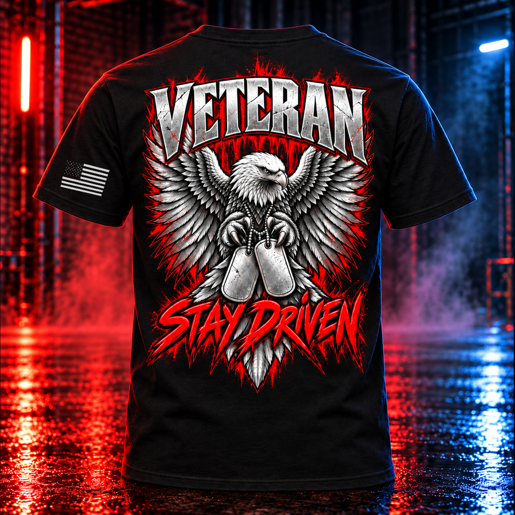 A black short-sleeve graphic T-shirt shown from the back, featuring a large eagle and crossed shotgun design with the words “VETERAN” at the top and “STAY DRIVEN” at the bottom in red and white. The shirt is photographed in a dramatic red-and-blue lit industrial setting with wet reflections on the floor.