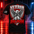 A black short-sleeve graphic T-shirt shown from the back, featuring a large eagle and crossed shotgun design with the words “VETERAN” at the top and “STAY DRIVEN” at the bottom in red and white. The shirt is photographed in a dramatic red-and-blue lit industrial setting with wet reflections on the floor.