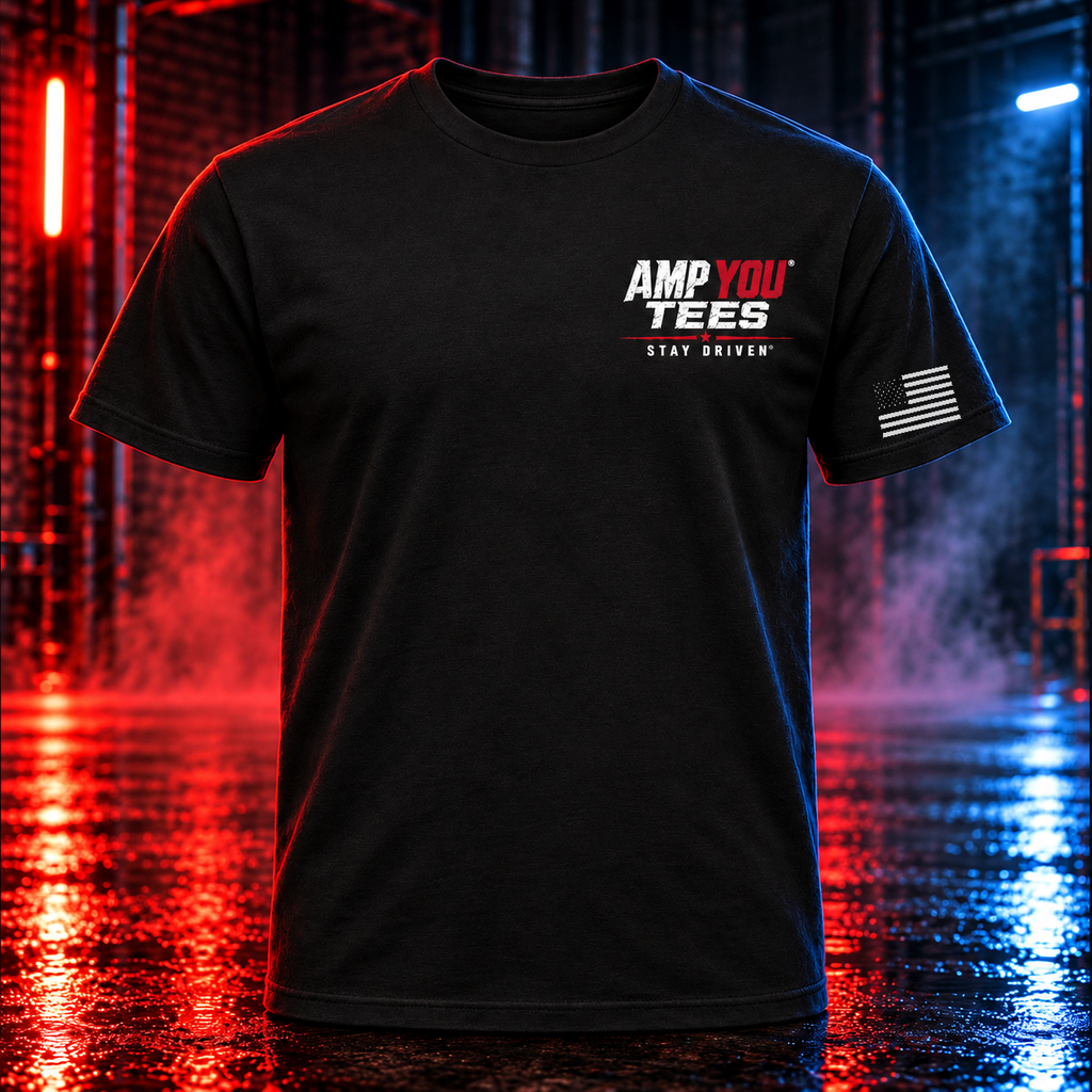 A black short-sleeve t-shirt with 'AMP YOU TEES' and 'STAY DRIVEN' printed on the chest, and a distressed American flag graphic on the sleeve, shown against a dark, wet, neon-lit industrial background.